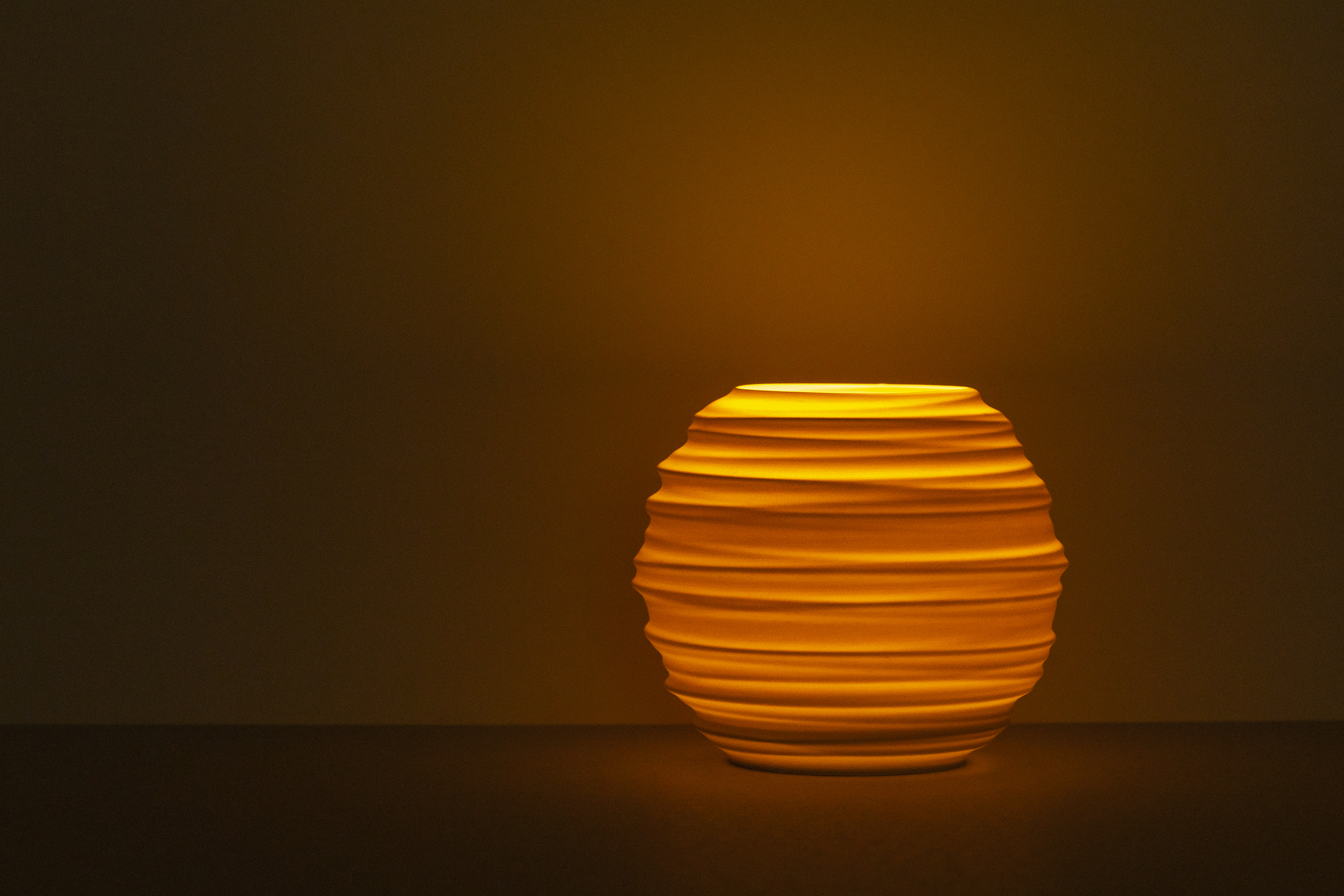 Decorative suggestion for Magma Shroud: Tealight holder inspired by magma's fiery embrace. Textured design casting elemental drama in shadows - variant Original (Creme and beige diffuse light), an original product by Cosmic Jubilee (1 of 3) - presented as a tealight, switched on.